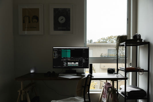 A modern, minimalist home office setup featuring a wooden desk with a monitor displaying a video editing program, a backlit salt lamp, and a side shelf with plants and cameras near a large window.
