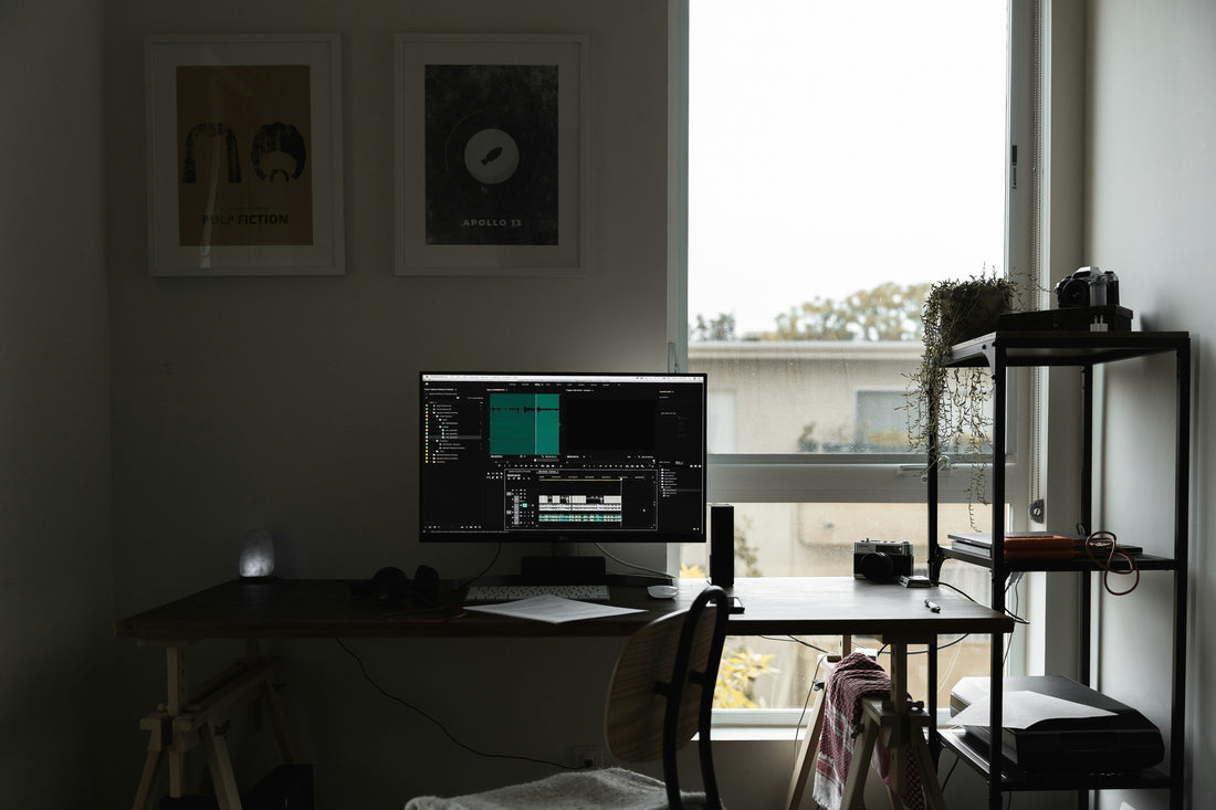 A modern, minimalist home office setup featuring a wooden desk with a monitor displaying a video editing program, a backlit salt lamp, and a side shelf with plants and cameras near a large window.